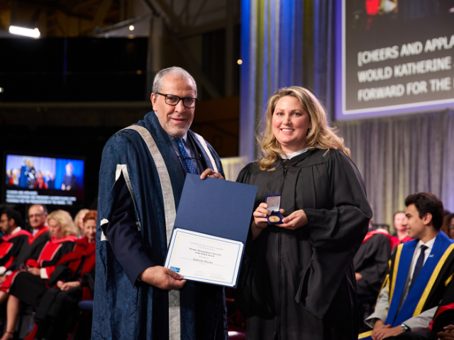 A woman in a graduation gown receives an award from a man in academic regalia on stage.