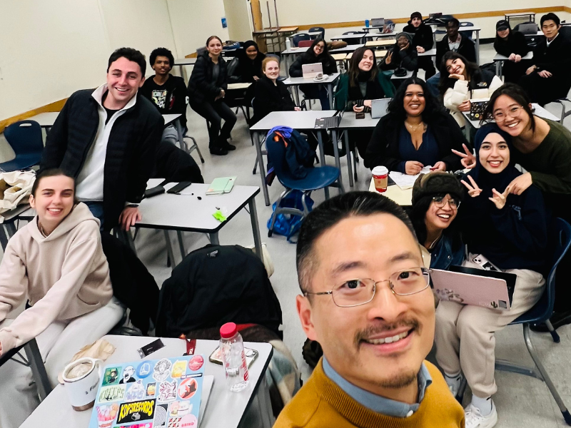 A diverse group of smiling students and a teacher pose in a classroom.
