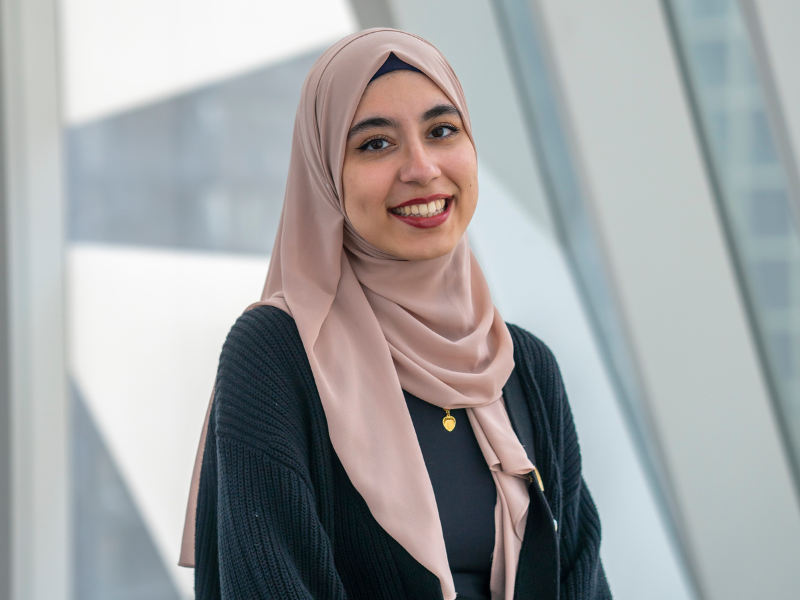 Woman in a light pink hijab and black sweater smiles at the camera.
