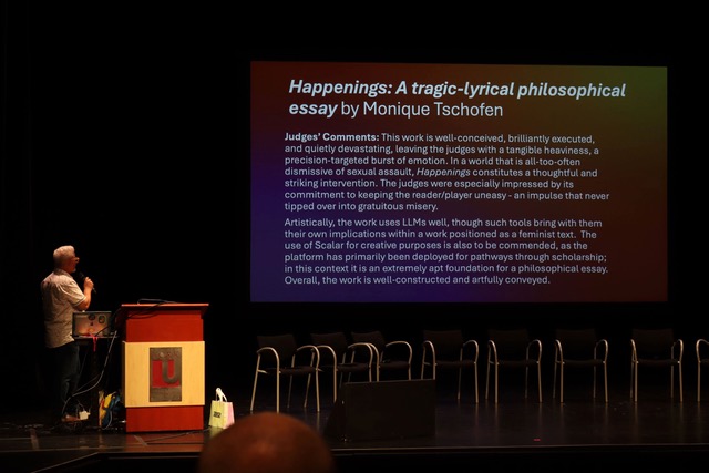 A person stands on a stage next to a podium, presenting in front of a large screen displaying the title "Happenings: A tragic-lyrical philosophical essay by Monique Tschofen"