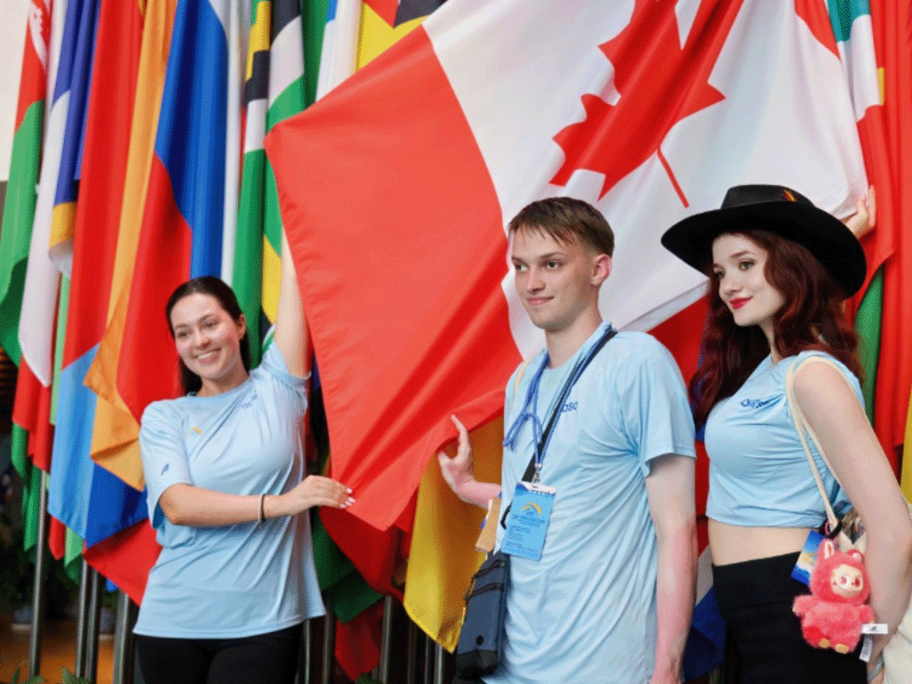 Three smiling people in blue shirts stand smiling in front of international flags, holding up the Canadian flag. 