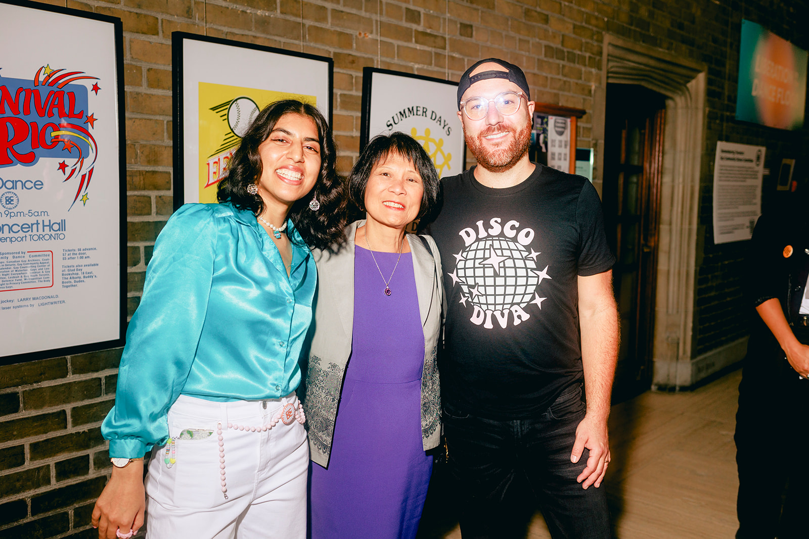 Two women and a man smiling at the camera, in the background are handbill posters.