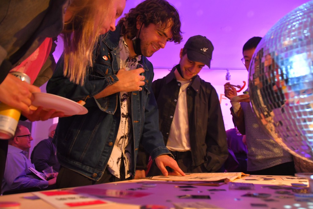 Group of men smiling, looking down at an open page of images. One man is pointing at a photo.