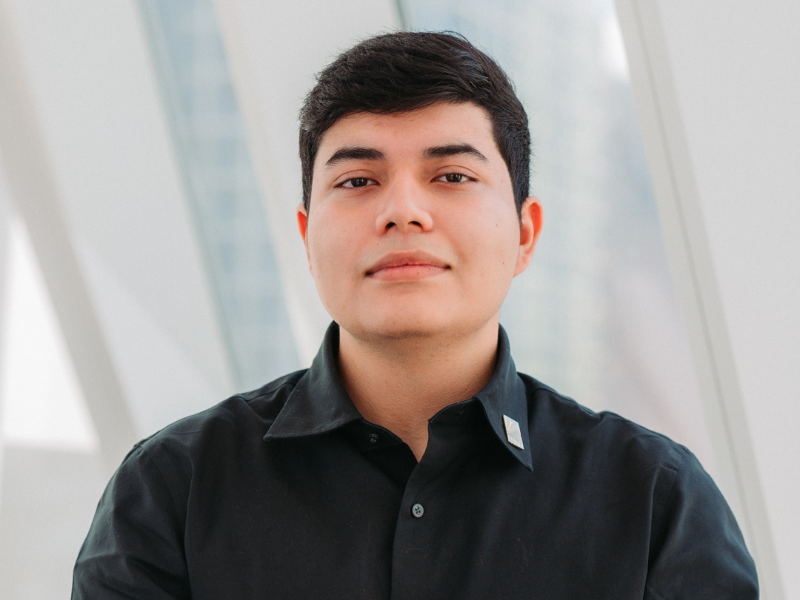  A man with short dark hair wearing a black button-up shirt poses confidently indoors, with a blurred background. 