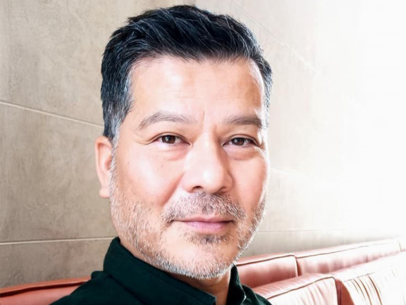 A man with short dark hair and a light beard wearing a black shirt smiles subtly.