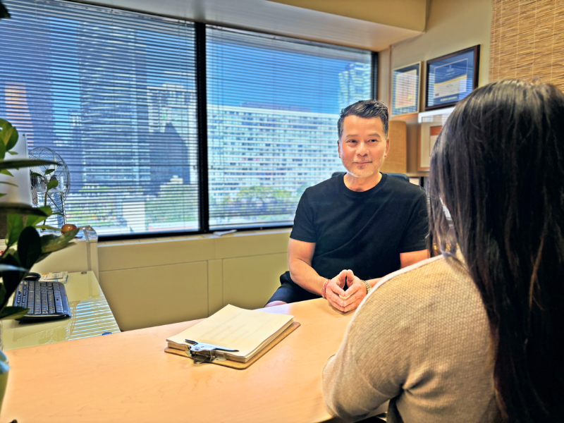 Sonny, in a black shirt, sits across from a student in an office with large windows and city views. 