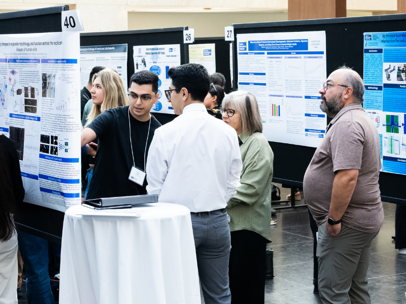 Daniel points at his poster. They are engaged in discussion, surrounded by research posters displaying charts and images.