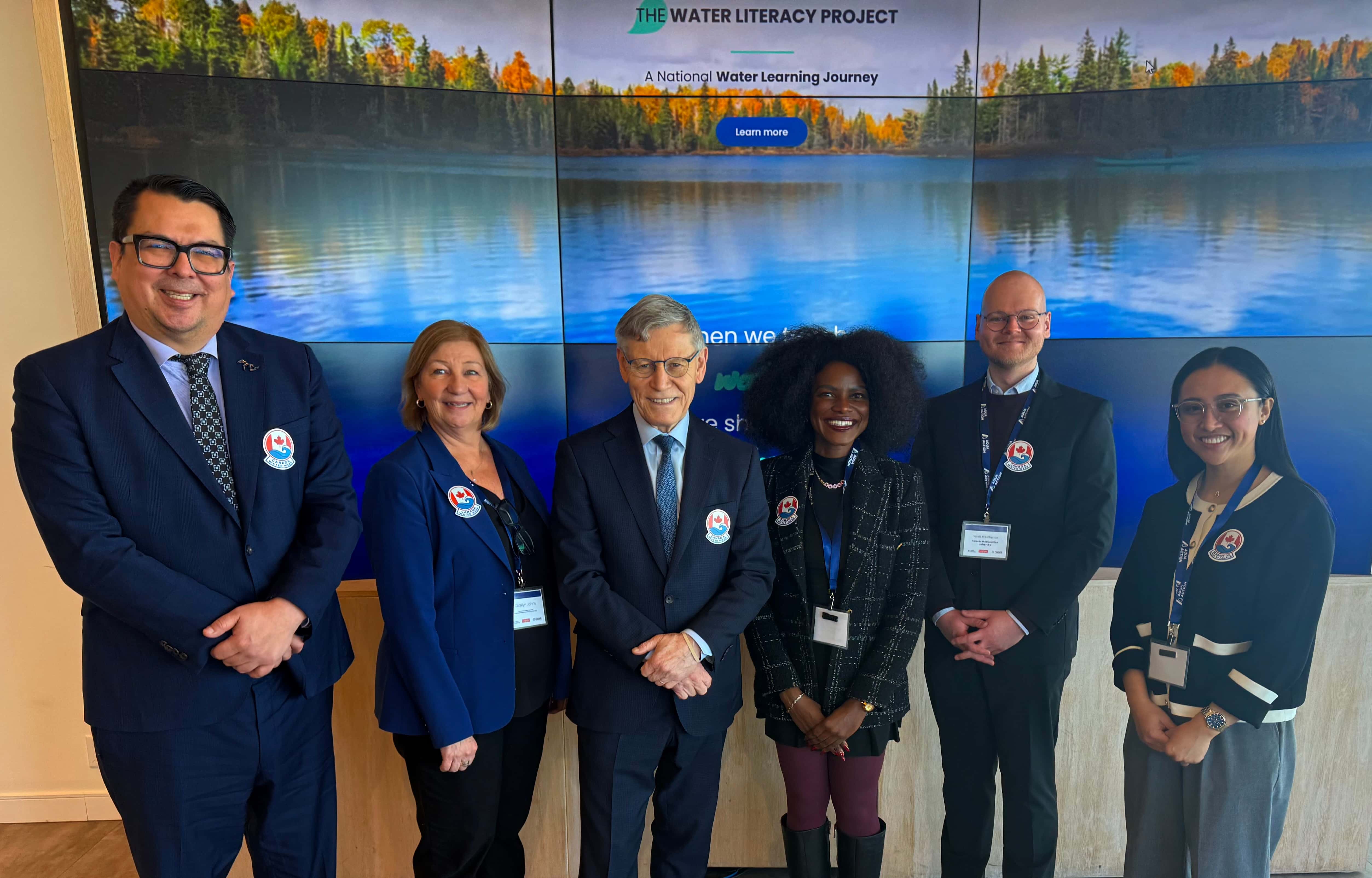 A group of 6 individuals are posed in a row smiling at the camera. They are all wears formal attire and are looking at the camera. They are wearing a specific Canada Water Week logo on their pins which are showing clearly. 