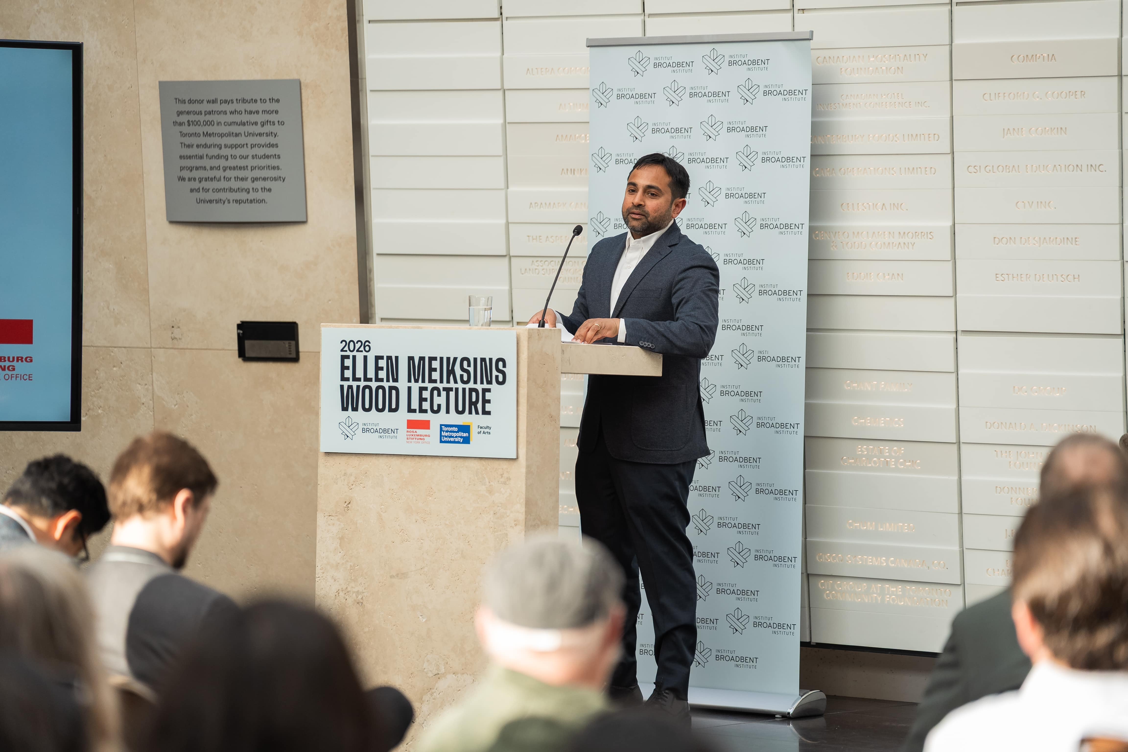 Bhaskar Sunkara is the figure centrally placed in the image with audience members backs and heads in the foreground. Bhaskar is wearing a grey suit with a high collar white shirt underneath at a podium with the Ellen Meiskins Sign shown. 