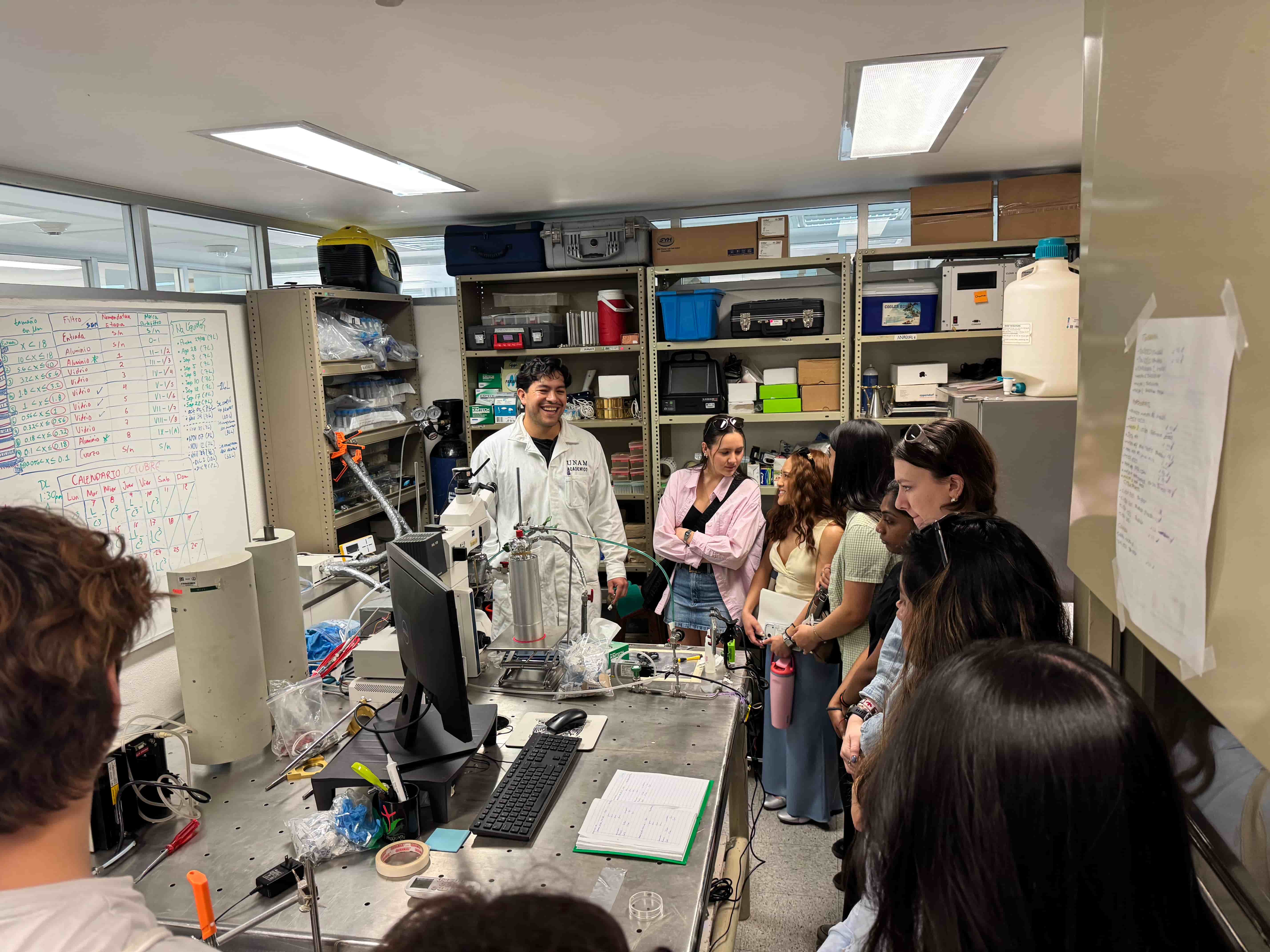 Students are in a small but unique laboratory observing a device in action. There is a lab coat person smiling and explaining the features of the machine. 
