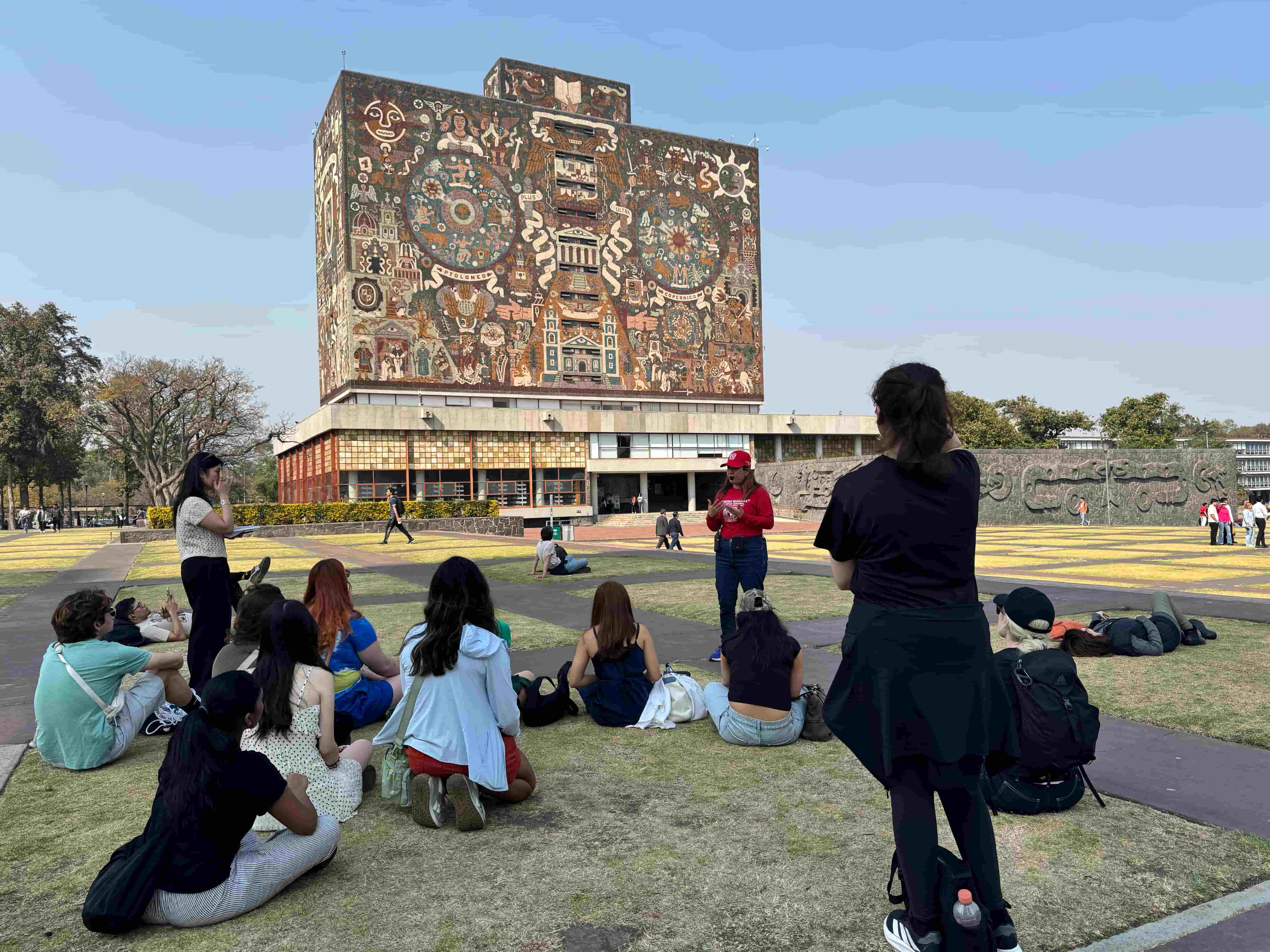 Students sitting down on shaded grass with a tour guide speaking. They are all listening attentively, facing away from the camera, while the tour guide speaks diligently. 