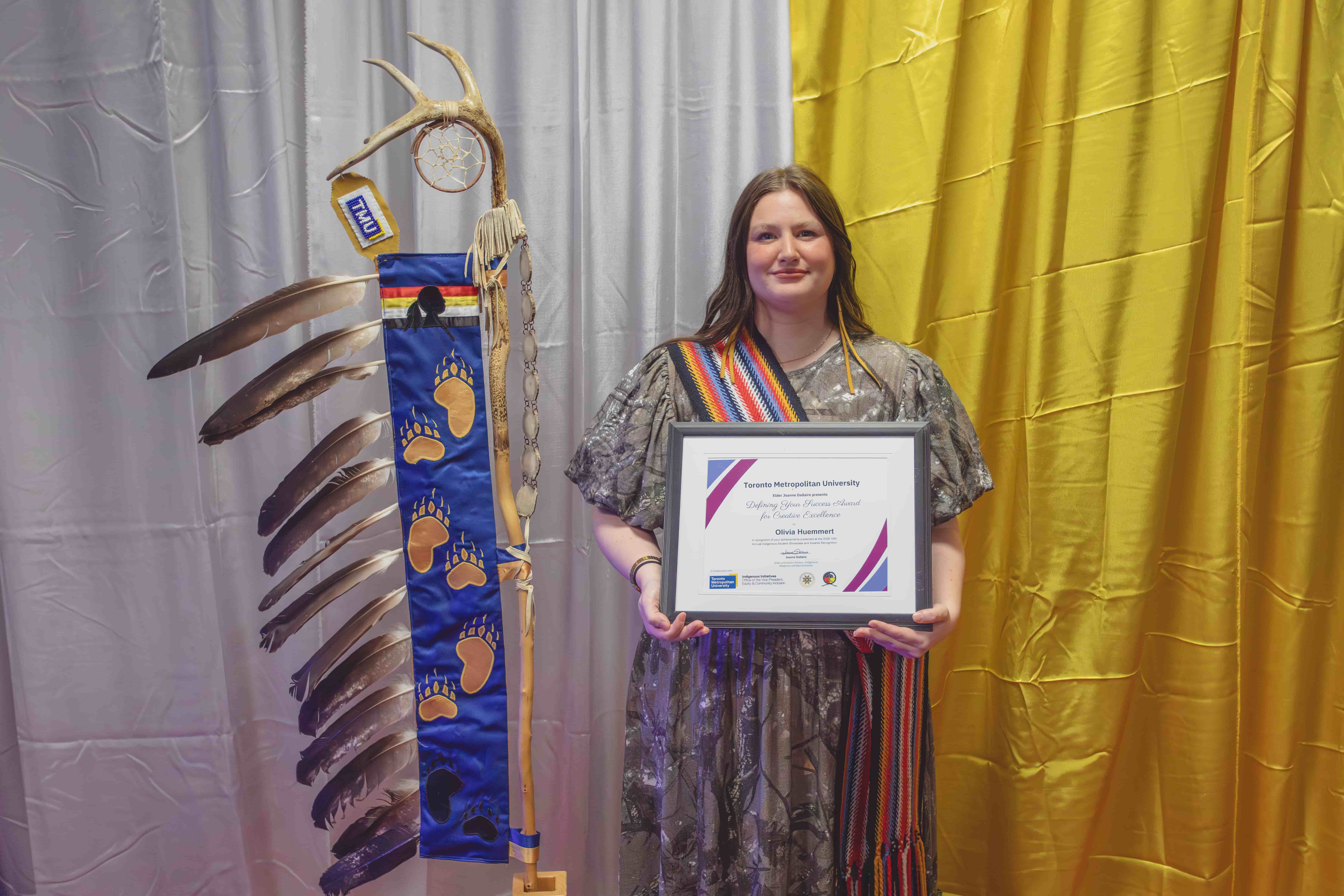 A student with an indigenous sassh around her shoulder towards her body is depicted holding an award that is framed behind a gold and silver background. She is standing next to a bone staff wth feathers and a small TMU flag.