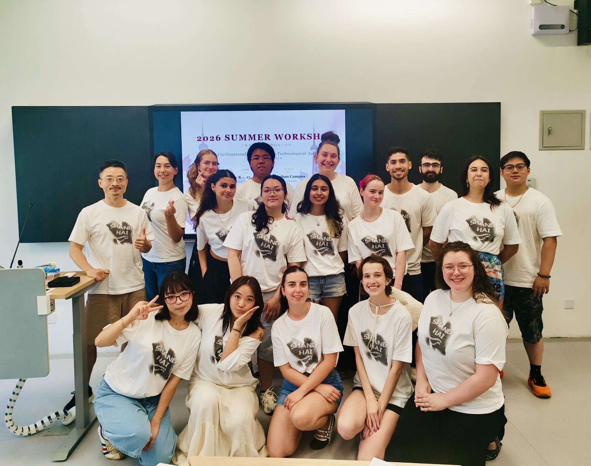A diverse group of students wearing the same t-shirt are posed infront of a screen in a classroom. They are all smiling for a group photo. The t-shirt is white and depicts the Shanghai University logo and their title. 