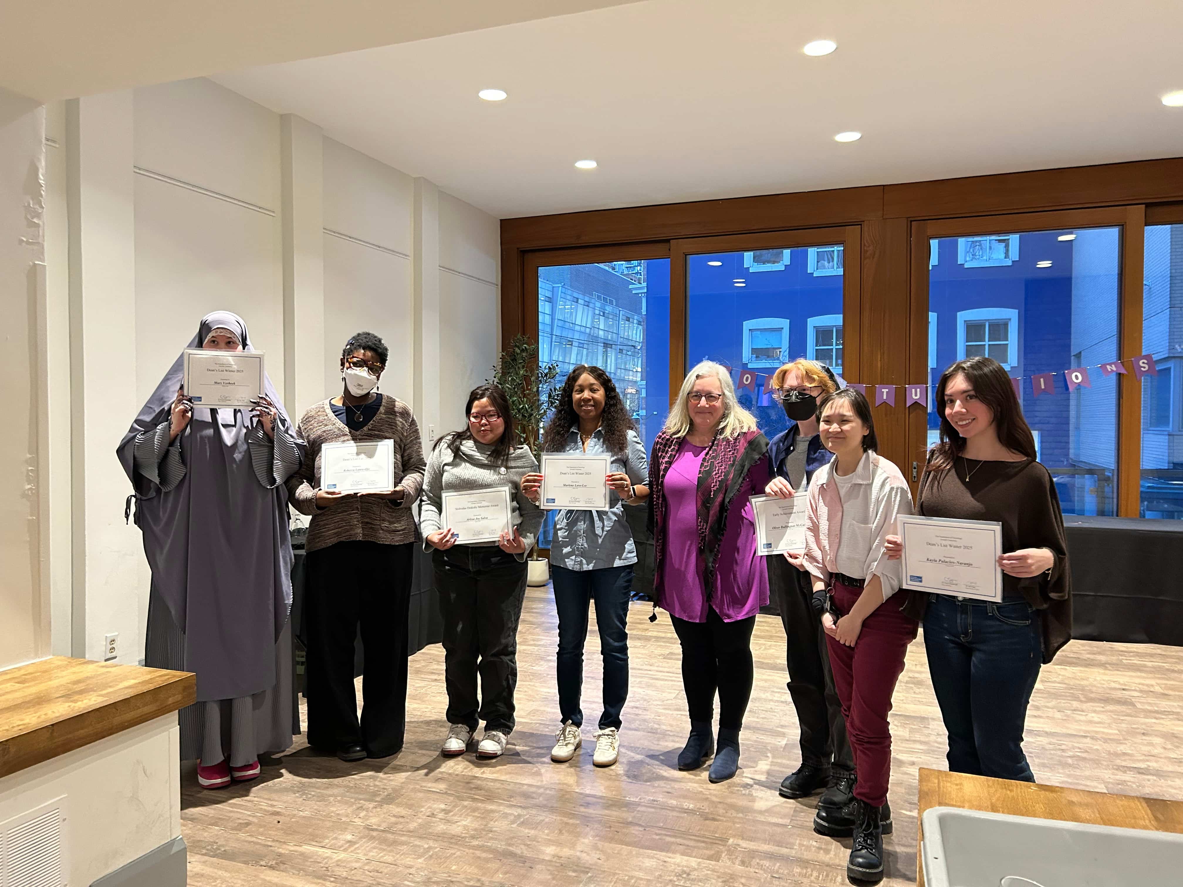 Students are shown with a professor, holding their awards in a wooden room. They are all smiling.
