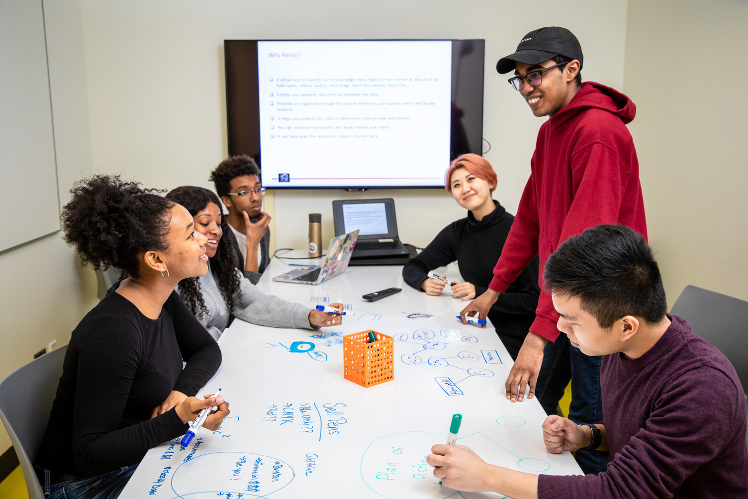 A group of students smiling in collaboration with each other, using whiteboard markers on a whiteboard table. They are in a library study room with a tv screen in the background.