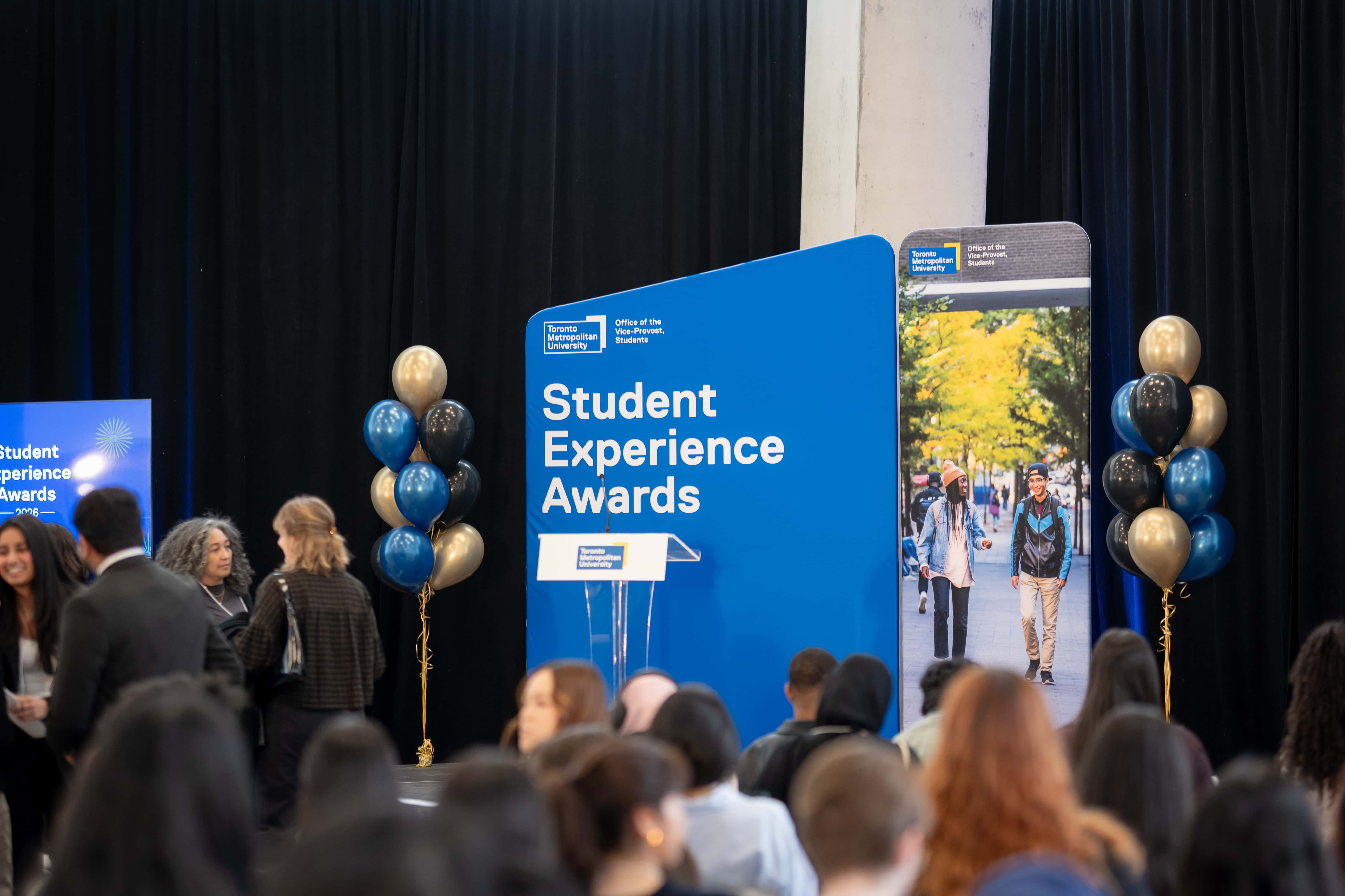 Podium of the Student Experience Awards is displayed. It features balloons in TMU colours, and students in the foreground. 