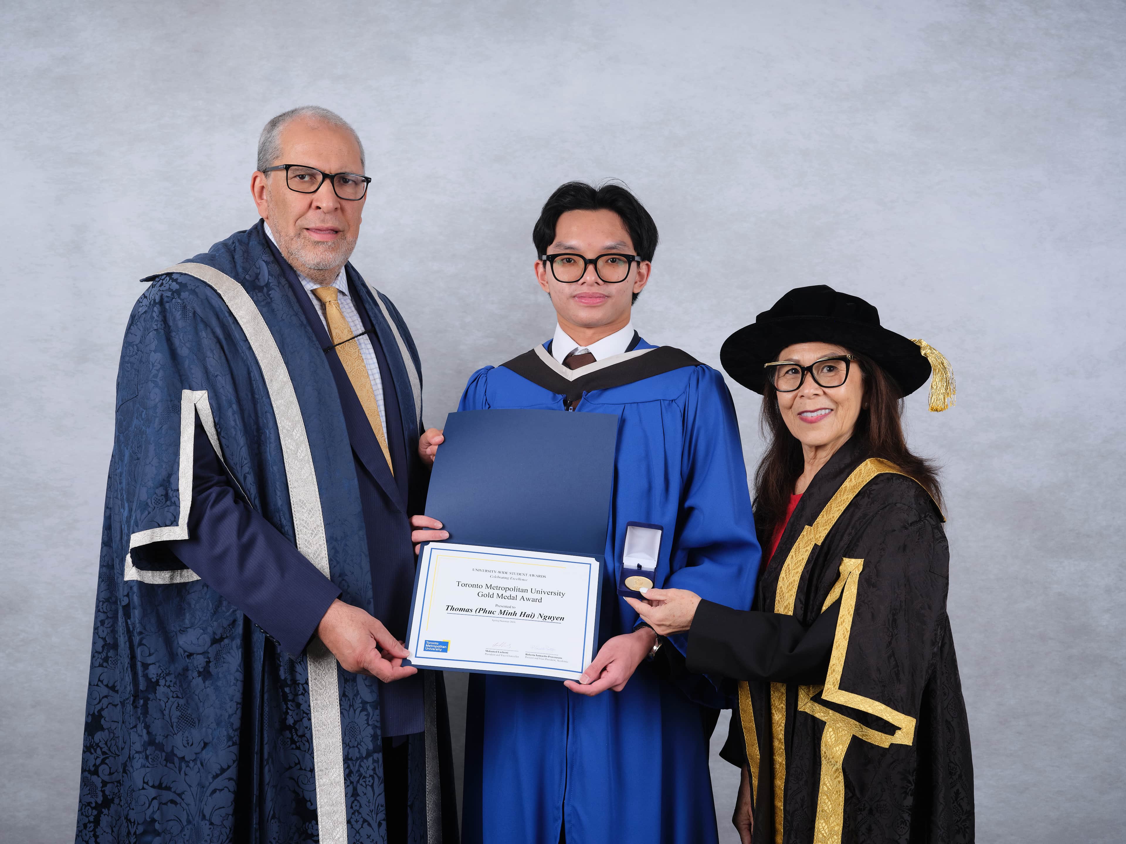 Thomas Nguyen, the winner, in a blue gown as graduation regalia. President Lachemi is wearing a blue gown as well as part of his attire, and the gown has white stripes on both sides. Chancellor Chang is wearing a black gown with a thick gold lining. She is also wearing a big black hat and red underneath the gown. 