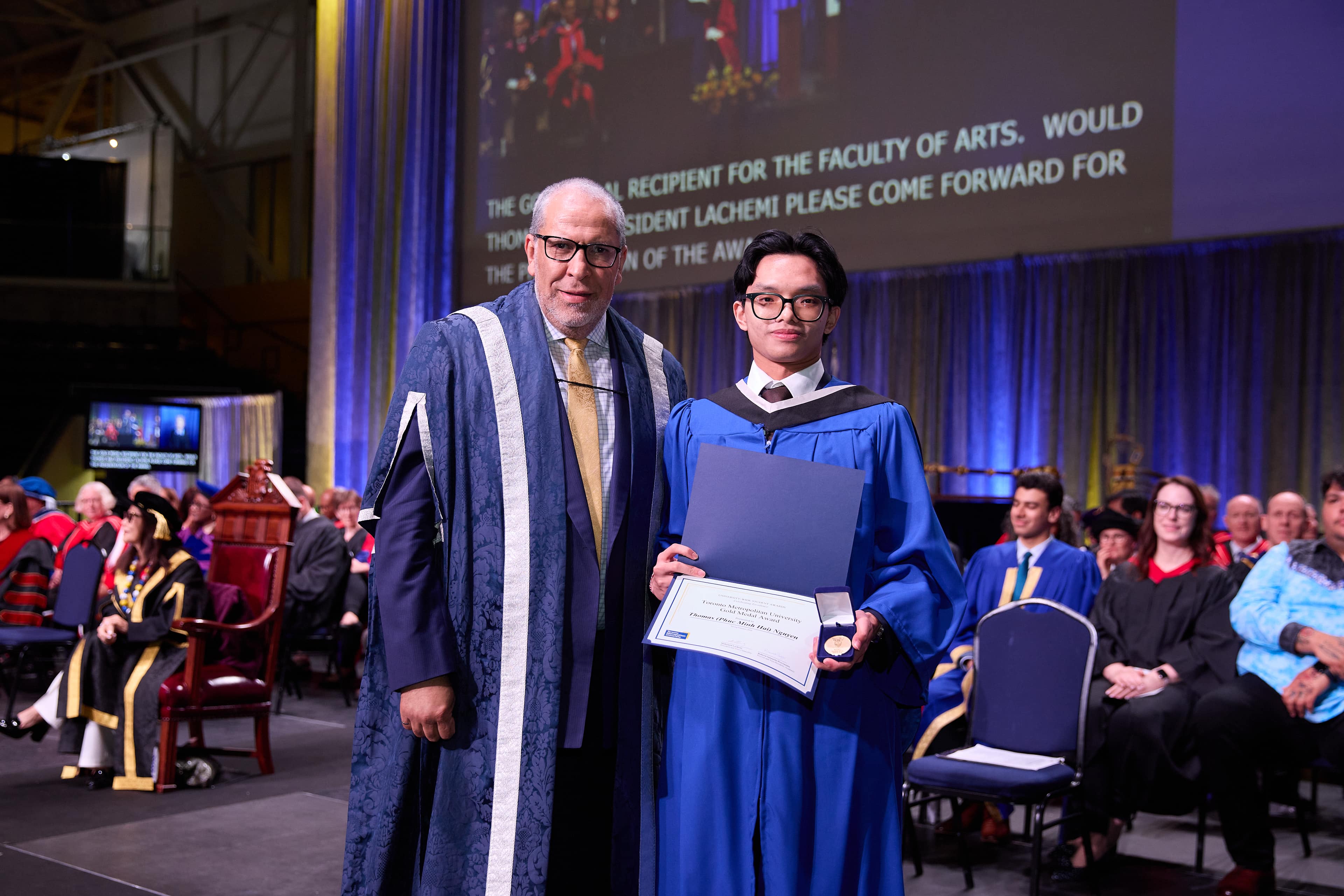 Thomas Nguyen in graduation regalia that is a blue gown with President Lachemi who is also wearing a blue gown with white stripes and a gold tie.