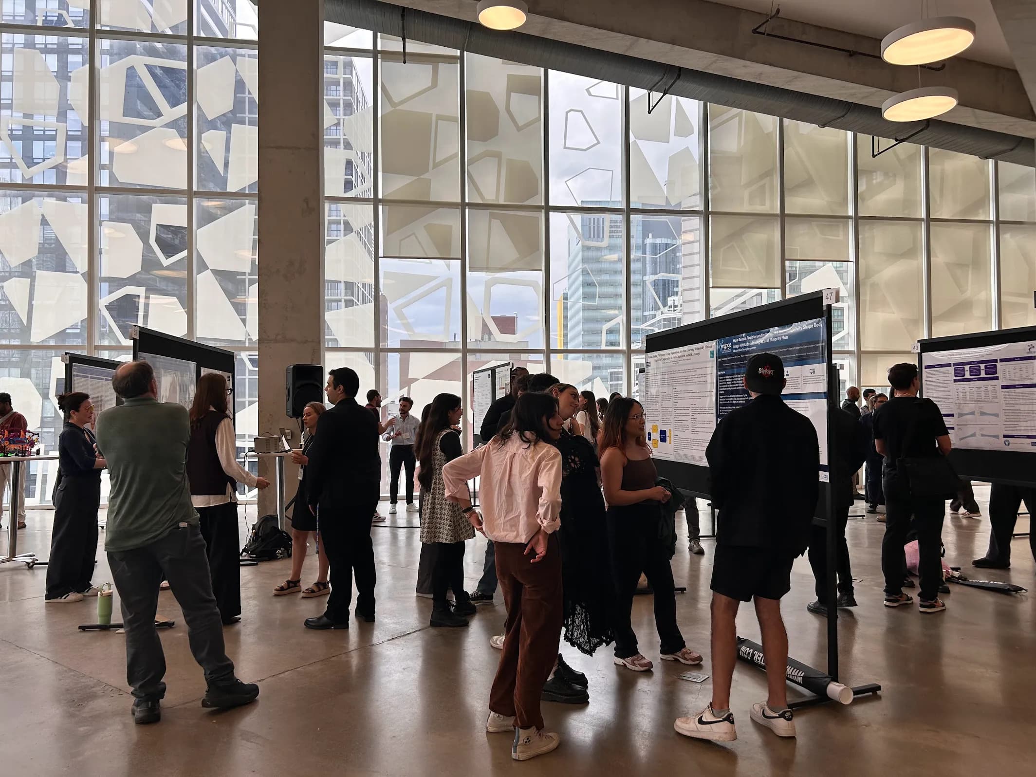A wide shot of the room where the showcase was held. There are small groups of diverse individuals listening attentively to students presenting in front of their poster. The background is a glass wall from the 8th floor of the SLC 