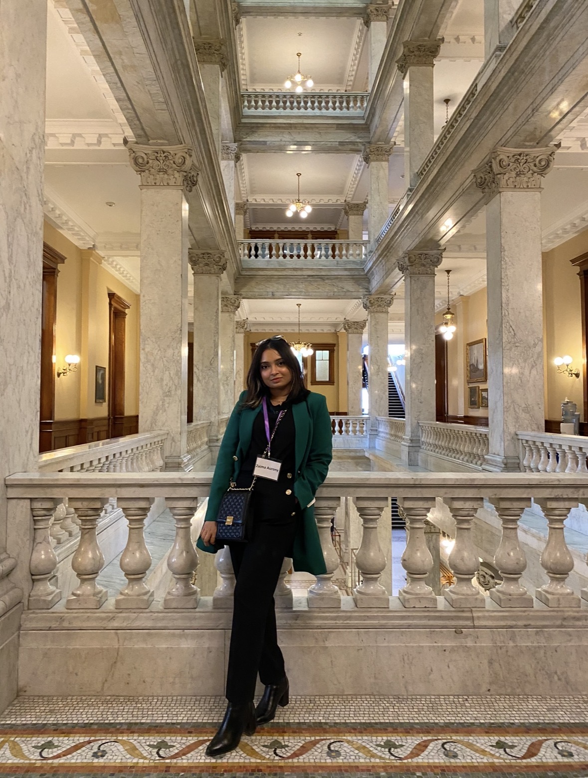 A women in a green blazer and formal attire is standing in a grand hall with mutliple floors. She is standing on beautiful tiled marble and in a space that is beautifully light with warm lighting