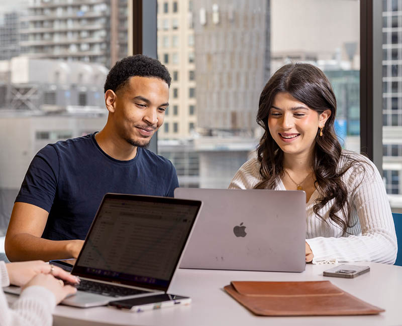 Two individuals sit at a table with laptops, smiling while working together. 