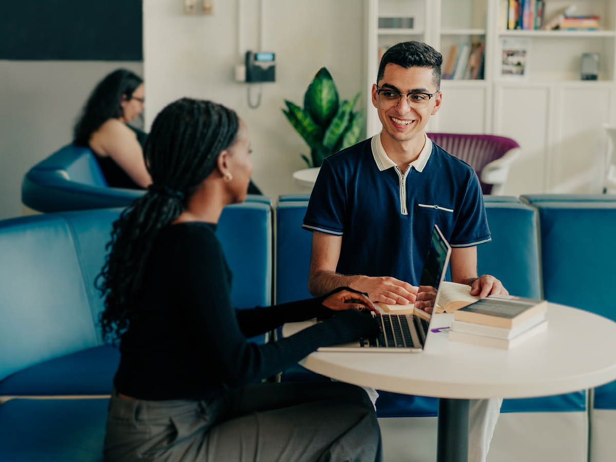 Two people smiling and talking at a table with a laptop and books in a modern lounge.