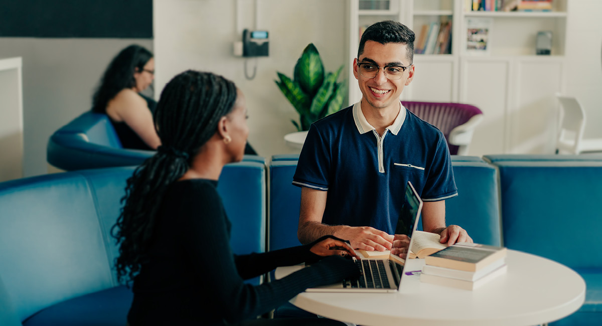 Two people smiling and talking at a table with a laptop and books in a modern lounge.