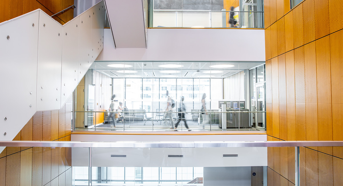 People walking through a bright modern office building with glass walls and wooden panel accents.