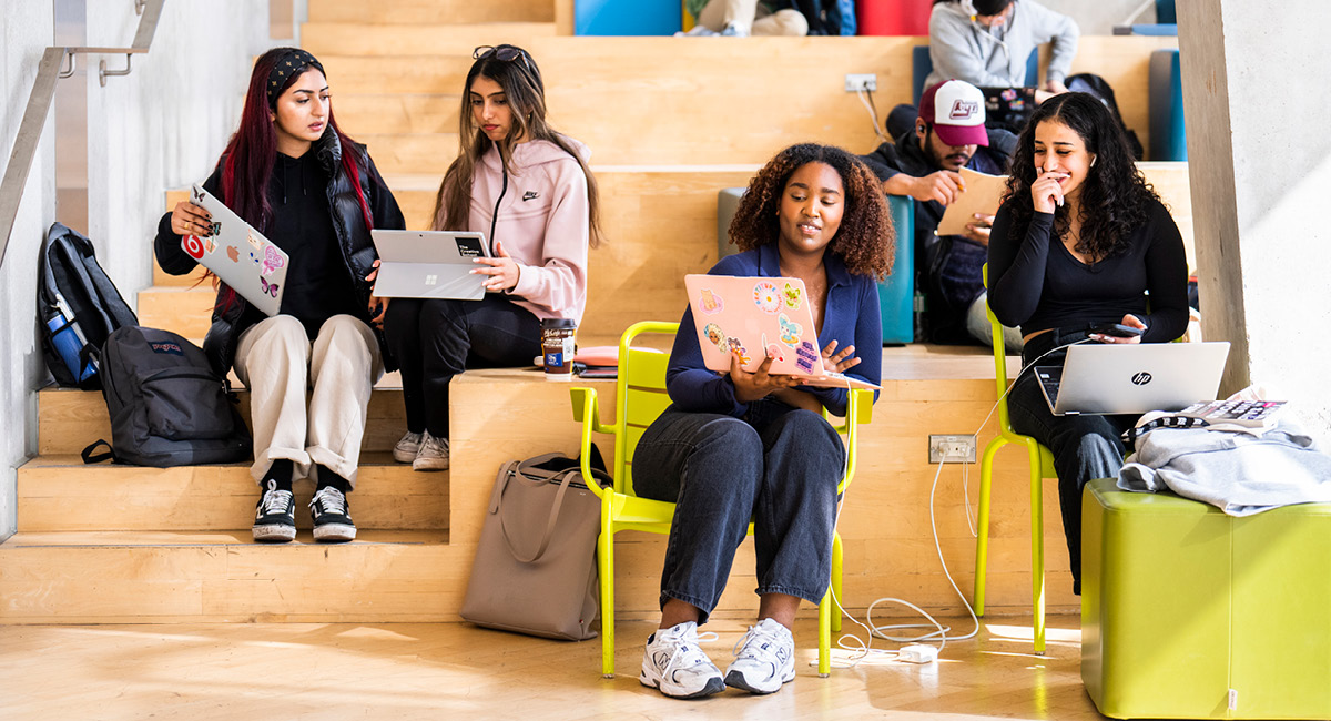 Four students sit on indoor steps with laptops, talking, backpacks, and charging cables nearby.