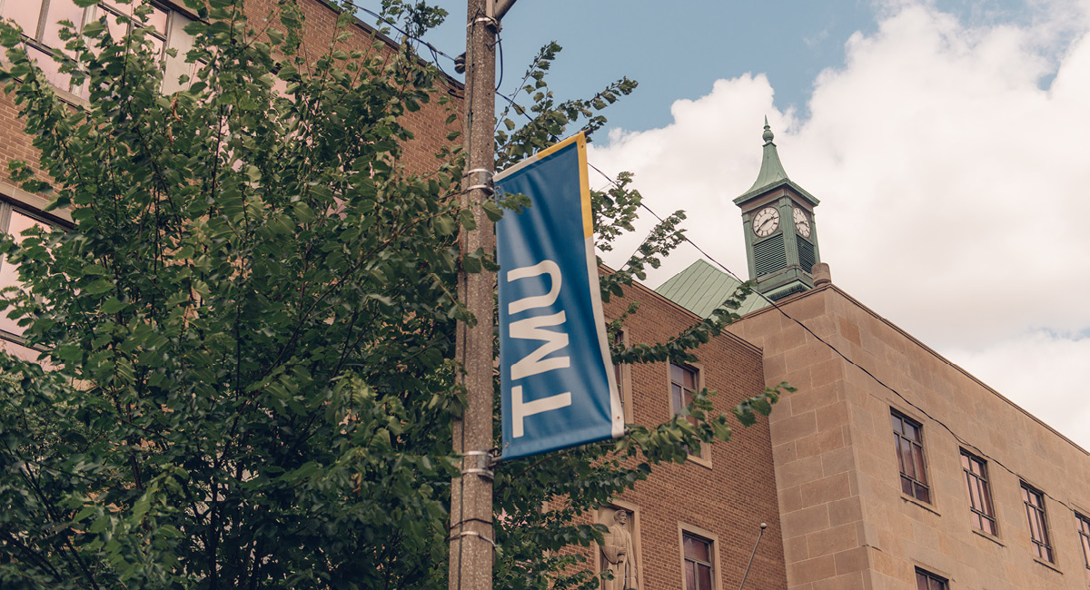 A TMU banner hangs on a pole beside a brick building with a green clock tower.