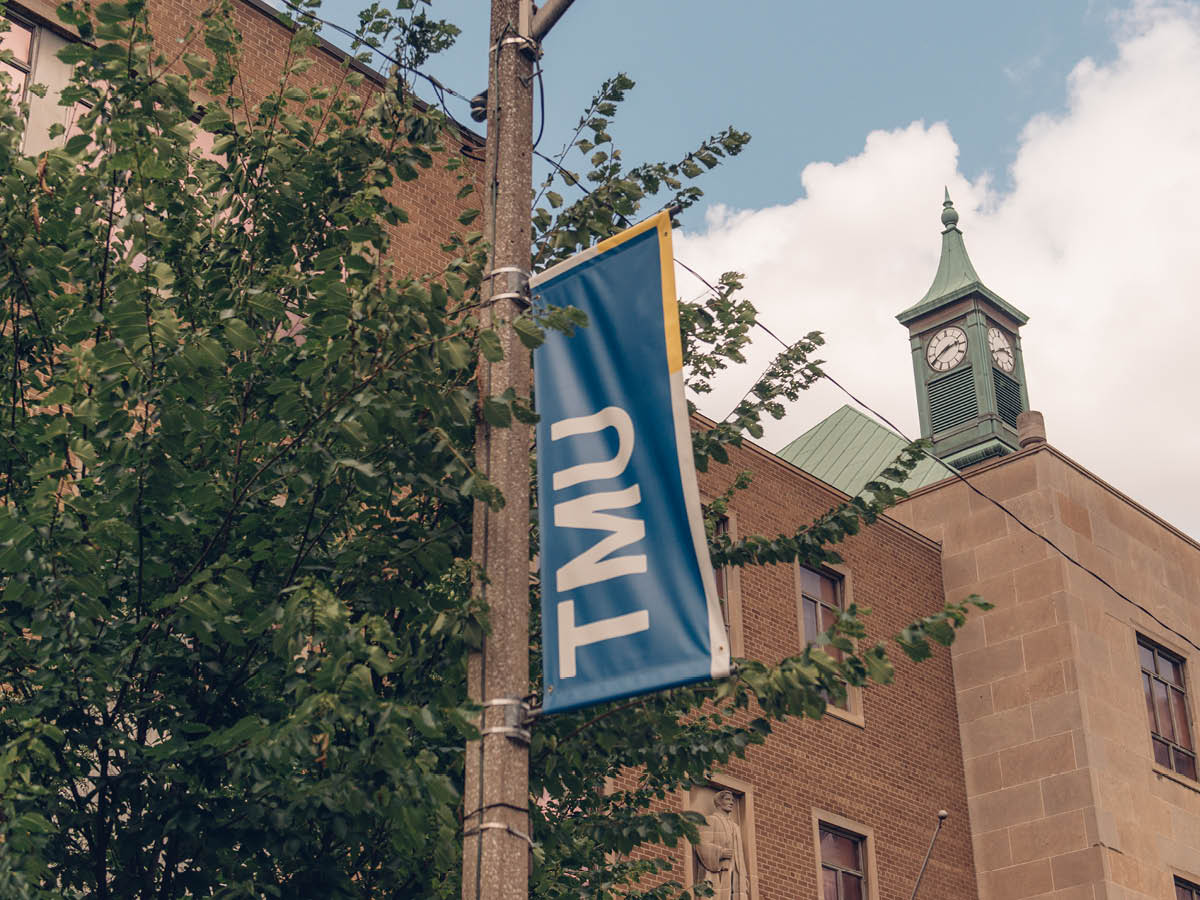 A TMU banner hangs on a pole beside a brick building with a green clock tower.