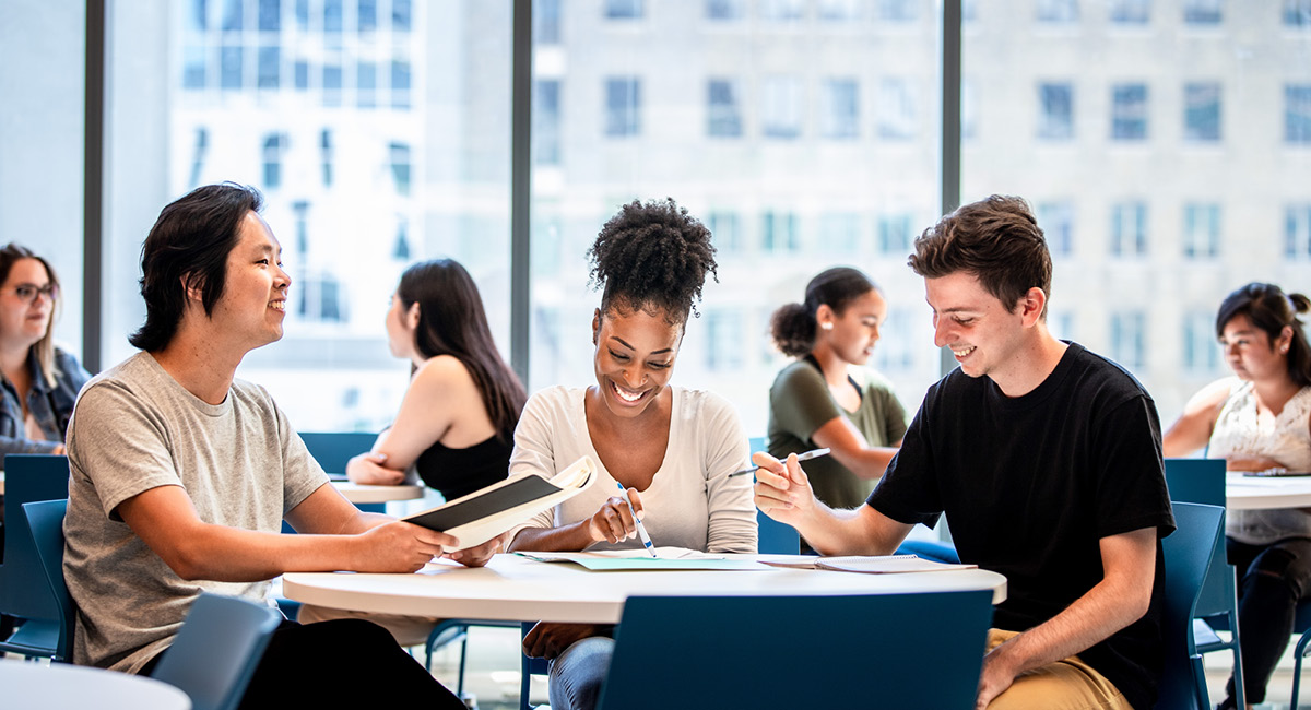 Three students study and smile at a table in a bright classroom with large windows.