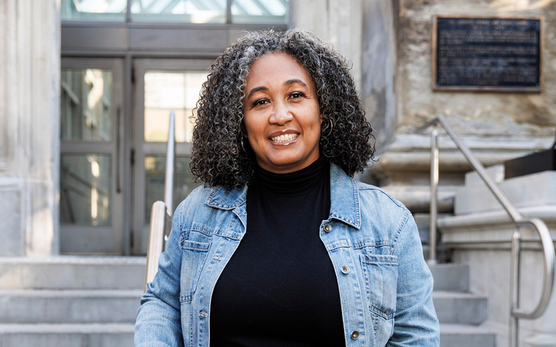 A woman with curly gray hair smiles warmly, wearing a denim jacket and black turtleneck on stone steps.
