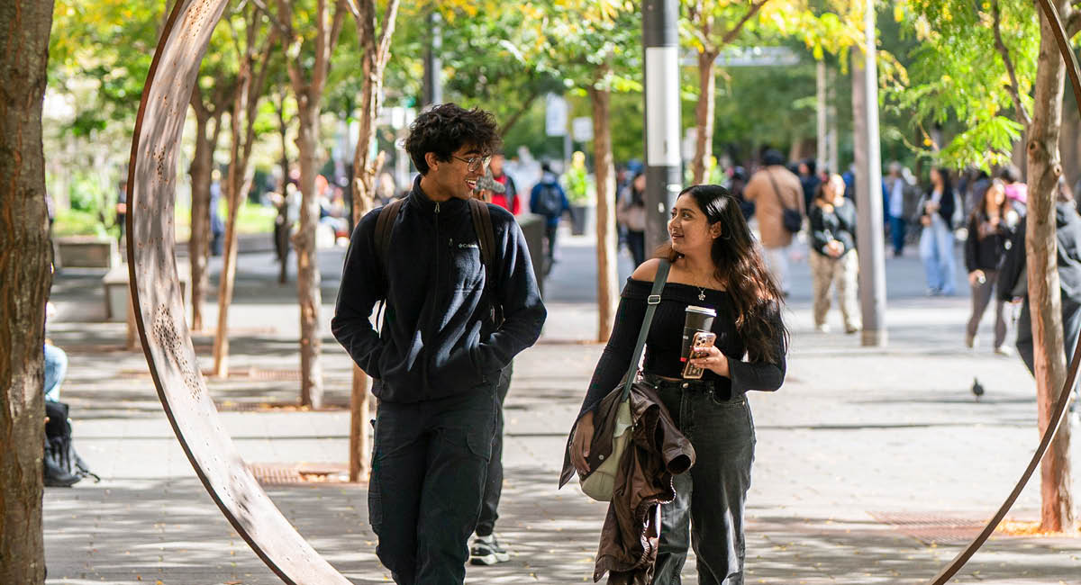 Two people walk and chat through park, framed by circular sculpture; trees and crowd create lively scene.