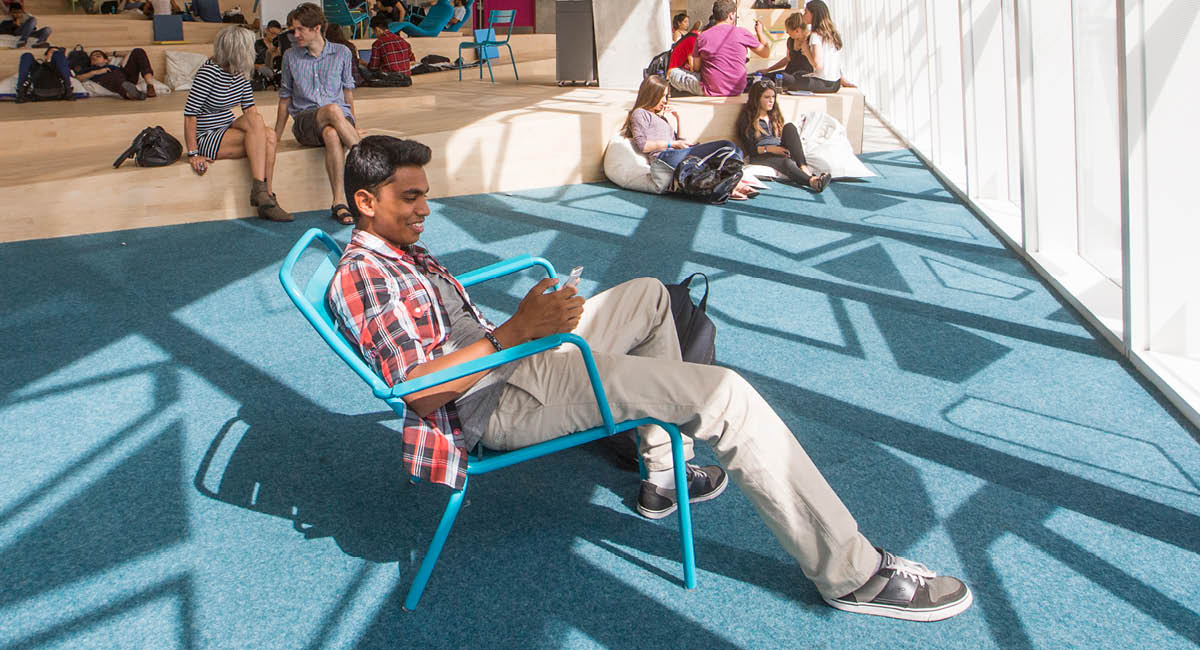 Student in red plaid shirt writes in notebook on blue chair; others relax on tiered seating in bright room.