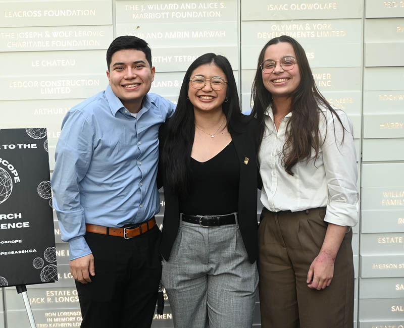 Three smiling people stand close together, dressed in business casual attire, in front of a wall with illuminated text.