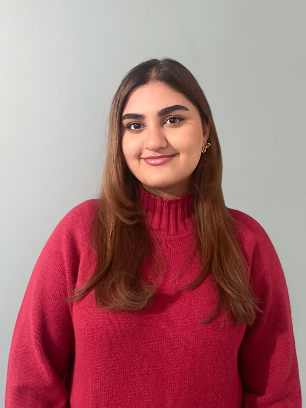 Smiling woman in red sweater with long brown hair and gold hoop earrings on light gray background.