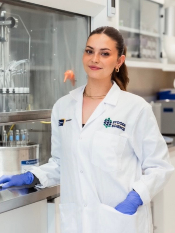 Woman in lab coat with tied-back hair and blue gloves stands in a lab with equipment behind her.