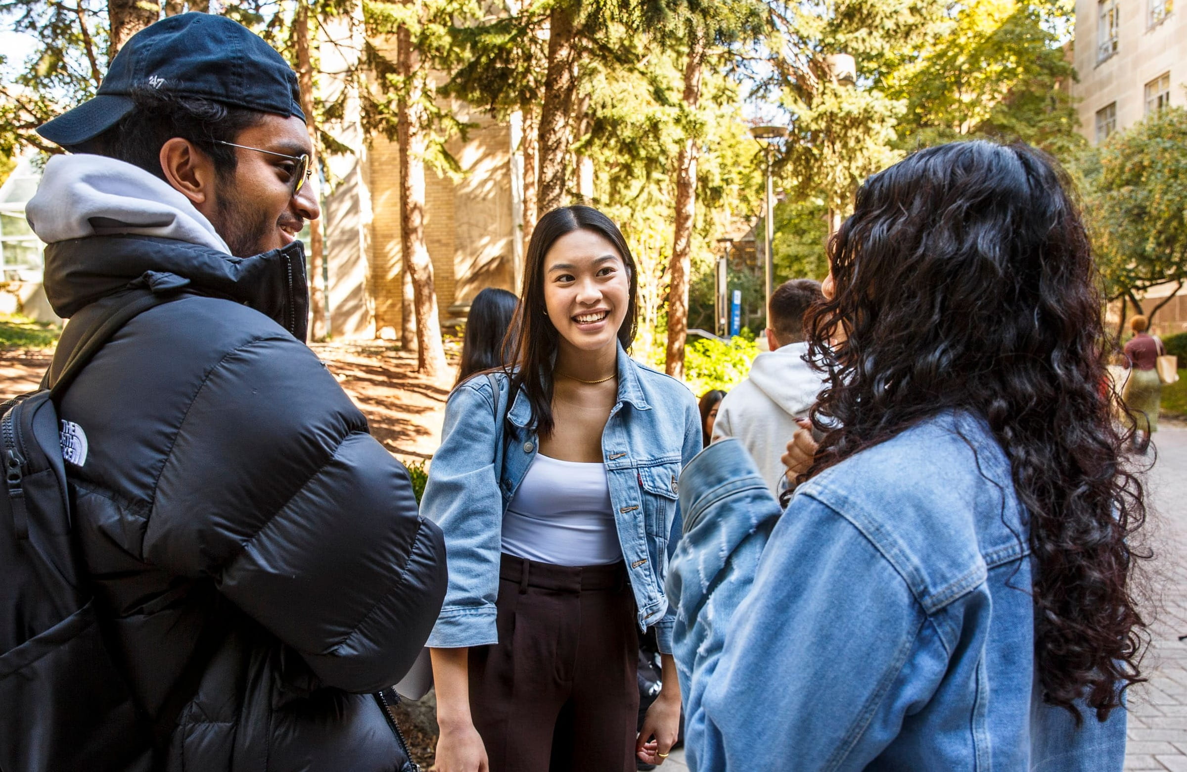 A diverse group of three students talking joyfully. One of the students is wearing a denim jacket and facing the other student and smiling. 