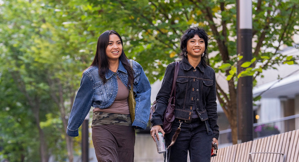 Two smiling people walk on tree-lined street, one in denim jacket, the other in black, relaxed and happy.