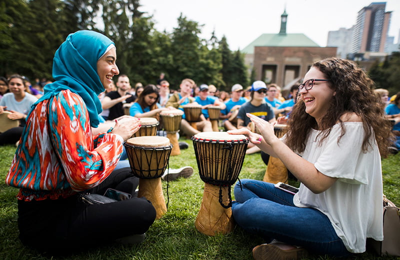 Diverse group drums in park; two women in front laugh, enjoying music in joyful, communal setting.