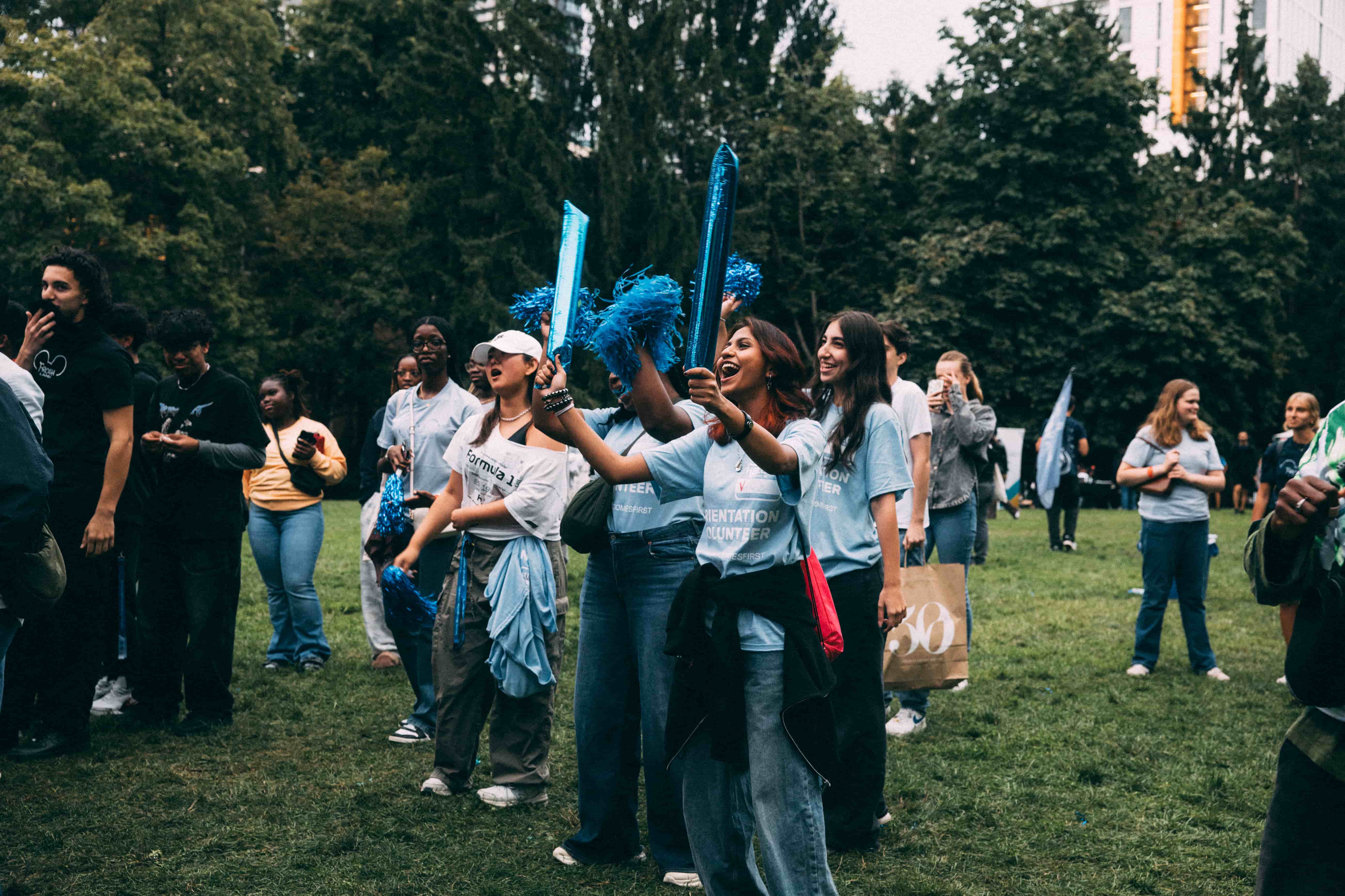 A group of students in light blue shirts highlighting their Orientation leader status looking left and cheering. One of them is holding two neon blue balloon sticks. They are surrounded by a group of new, diverse students and are on dark green grass. 