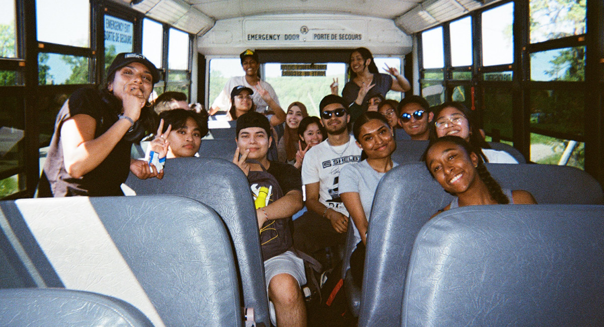 Joyful, diverse teens pose inside school bus, smiling and making peace signs in sunny, carefree atmosphere.
