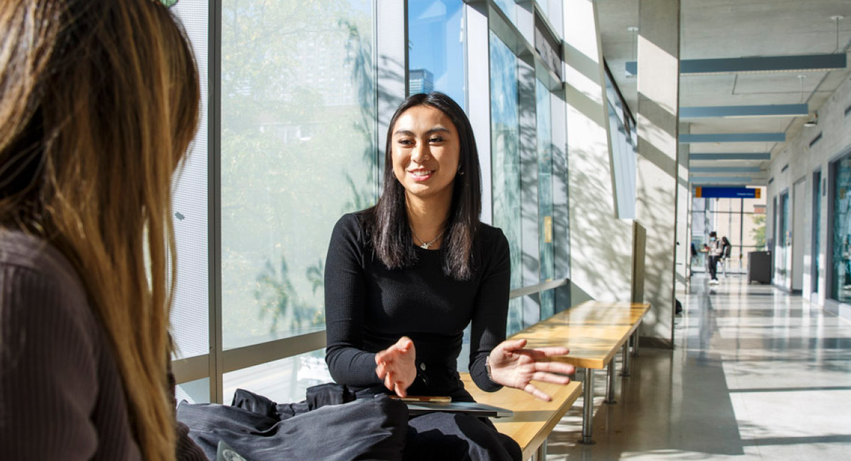 Woman in black talks energetically with another in sunlit hallway, sitting on wooden benches in friendly setting.