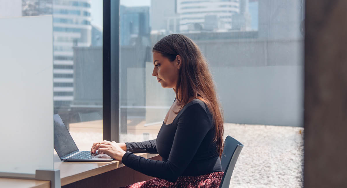 Woman with long hair types on laptop at desk in bright office with large windows showing cityscape.