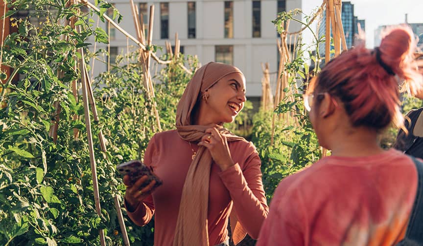 Two people smiling in a sunlit garden, one holding vegetables, with greenery and buildings behind.