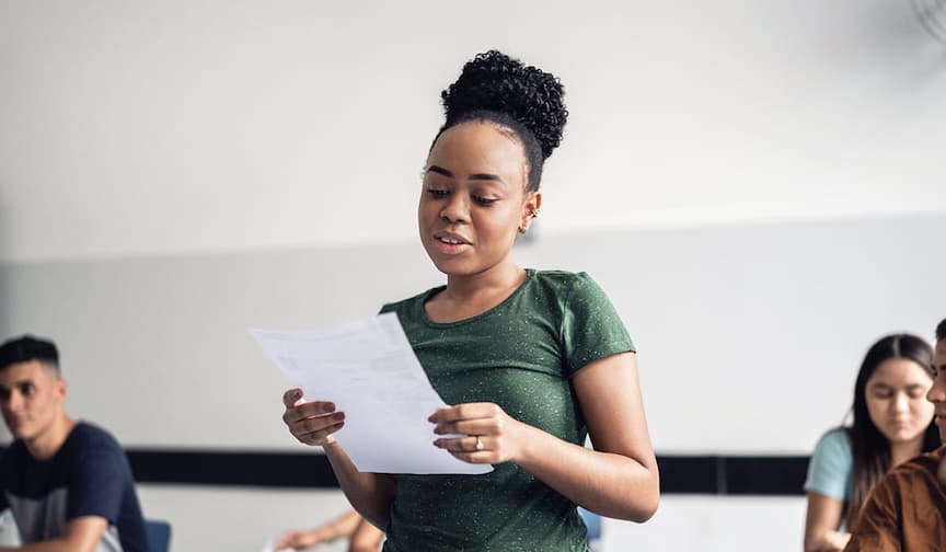 Young woman in green shirt reads confidently in a classroom, with peers seated and engaged behind.