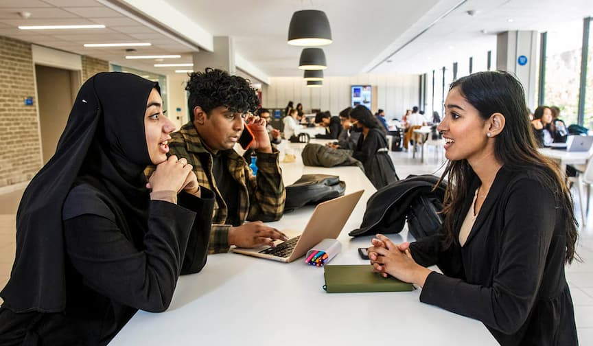 Three students sit at a white table in a modern study space, deep in a lively discussion.