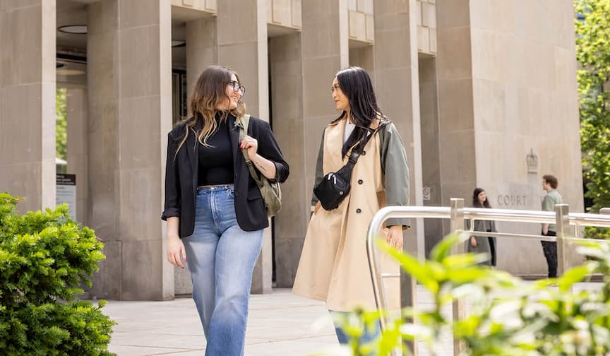 Two women walk and chat outside a modern building on a sunny day, surrounded by greenery.
