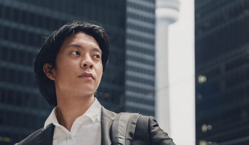A young man in a suit gazes upward in a cityscape, with skyscrapers and a tower in the background.